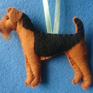 May include: A felt toy of a brown and black Airedale Terrier dog with a light blue ribbon hanging from its neck.
