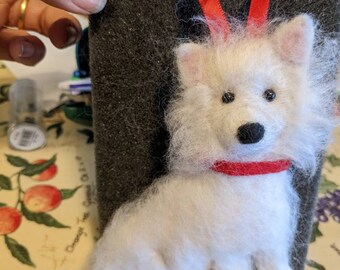 May include: A white felt dog ornament with a red collar, hanging from a red ribbon. The dog is sitting on a black foam board.