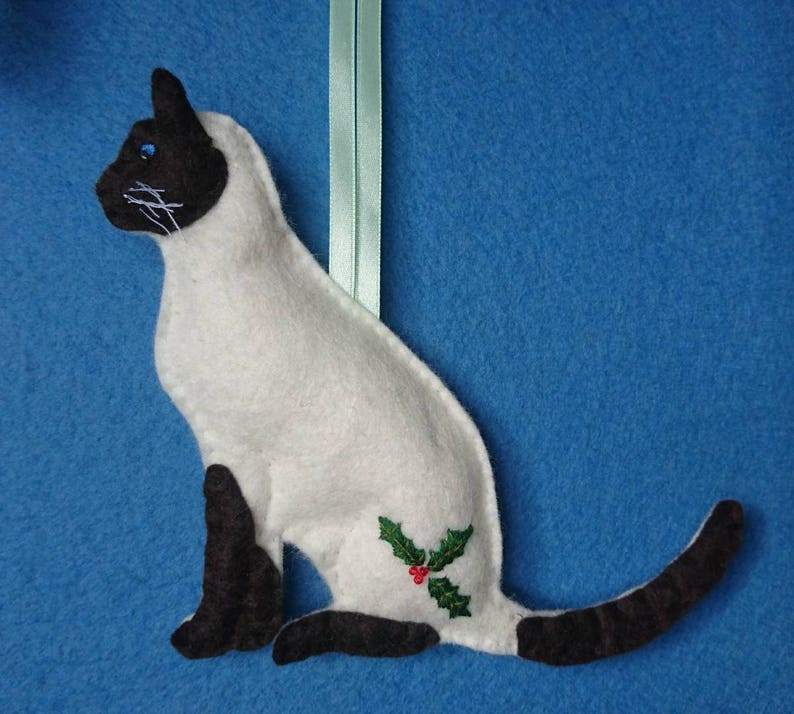 May include: A white felt Siamese cat ornament with black ears, a black tail, and blue eyes. The cat is sitting with its tail curled around its body. A small green holly sprig with a red berry is embroidered on the cat's side.