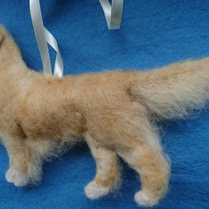 K&ouml;nnte beinhalten: Ein handgefilzter Golden Retriever Hundefiguren-Anh&auml;nger mit einem wei&szlig;en Band zum Aufh&auml;ngen. Der Hund steht auf einer blauen Oberfl&auml;che.