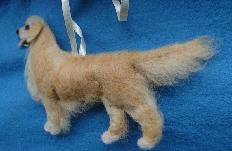 K&ouml;nnte beinhalten: Ein handgefilzter Golden Retriever Hundefiguren-Anh&auml;nger mit einem wei&szlig;en Band zum Aufh&auml;ngen. Der Hund steht auf einer blauen Oberfl&auml;che.