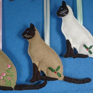 May include: Three felt Siamese cat ornaments, each with a different design. One cat has a holly design, one has a floral design, and one has a plain design. The cats are all sitting with their tails curled around their bodies.