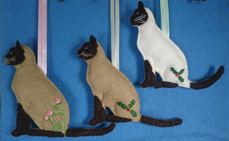 May include: Three felt Siamese cat ornaments, each with a different design. One cat has a holly design, one has a floral design, and one has a plain design. The cats are all sitting with their tails curled around their bodies.