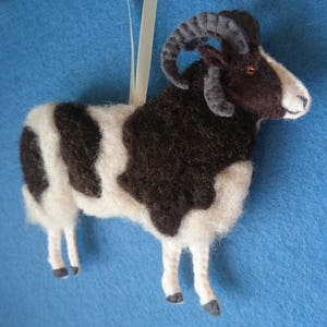 Puede incluir: Un adorno de lana fieltro a mano de una oveja blanco y negro con cuernos marrones. La oveja est&aacute; de pie sobre un fondo azul.