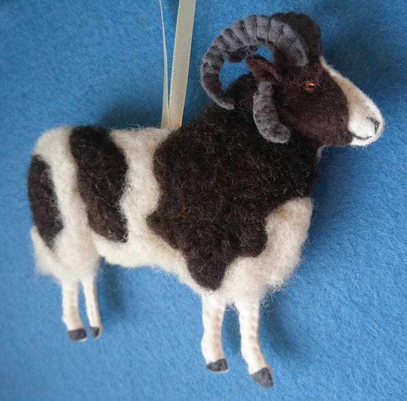 Puede incluir: Un adorno de lana fieltro a mano de una oveja blanco y negro con cuernos marrones. La oveja est&aacute; de pie sobre un fondo azul.