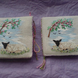 May include: Two small, square, beige fabric cushions with embroidered images of a white lamb sleeping under a tree with pink blossoms. The cushions have green and pink tassels.