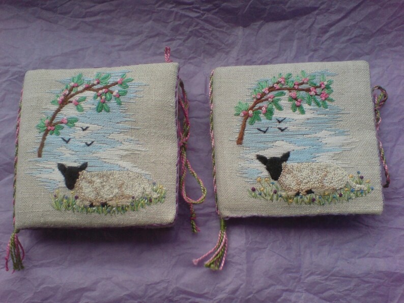 May include: Two small, square, beige fabric cushions with embroidered images of a white lamb sleeping under a tree with pink blossoms. The cushions have green and pink tassels.