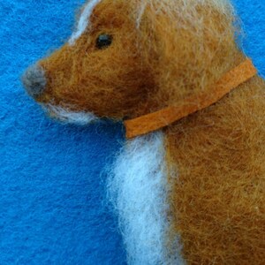 May include: A brown and white needle felted dog with an orange collar. The dog is facing to the left and has a white patch on its forehead.