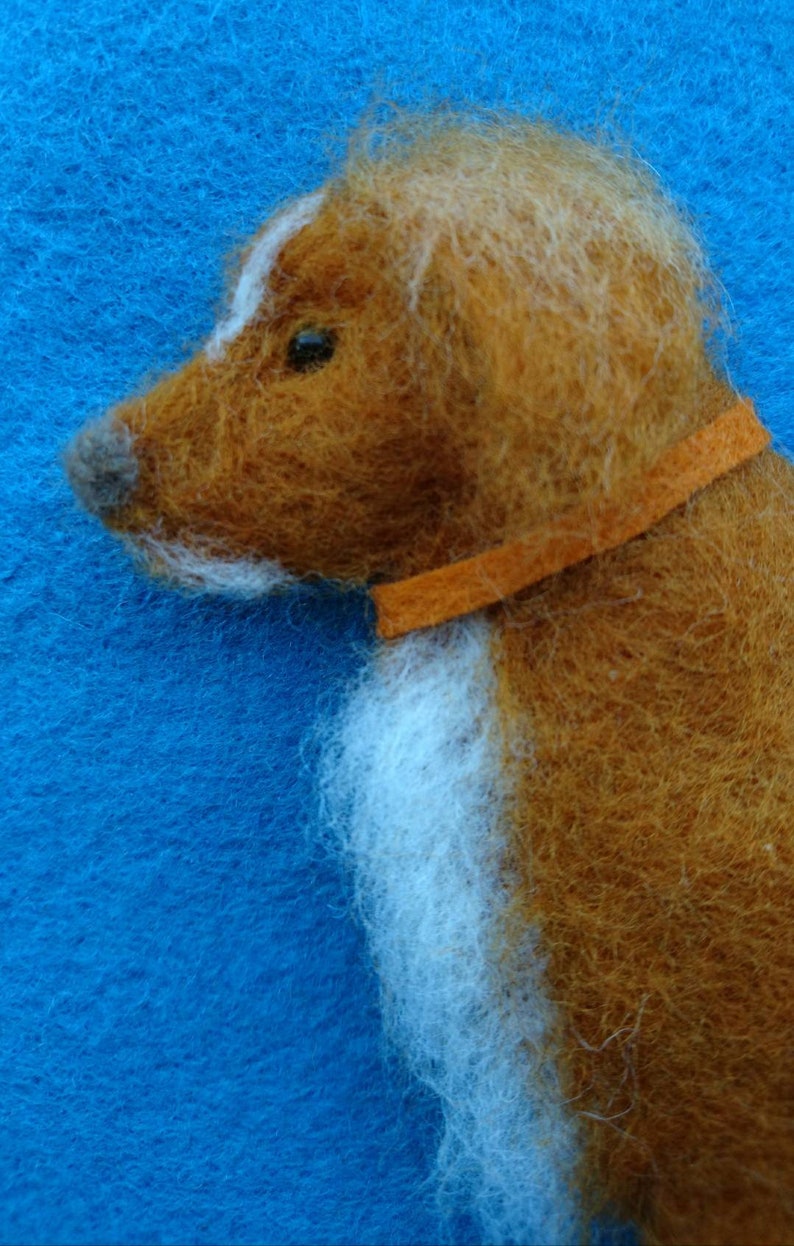 May include: A brown and white needle felted dog with an orange collar. The dog is facing to the left and has a white patch on its forehead.