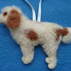 May include: A white and brown needle felted dog ornament. The dog is standing on all four legs and has a curly tail. The ornament is made of wool and is about 5 inches tall.