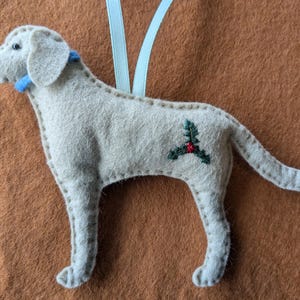 May include: A white felt dog ornament with a blue collar and a red and green holly design on its back. The ornament is hanging from a blue ribbon.