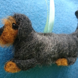 May include: A needle-felted dog ornament featuring a black and grey textured body with tan accents on the face, paws, and tail tip. The dog has a small black eye and a light green ribbon loop attached to the back for hanging purposes.