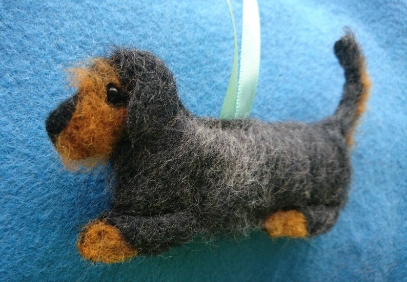 May include: A needle-felted dog ornament featuring a black and grey textured body with tan accents on the face, paws, and tail tip. The dog has a small black eye and a light green ribbon loop attached to the back for hanging purposes.