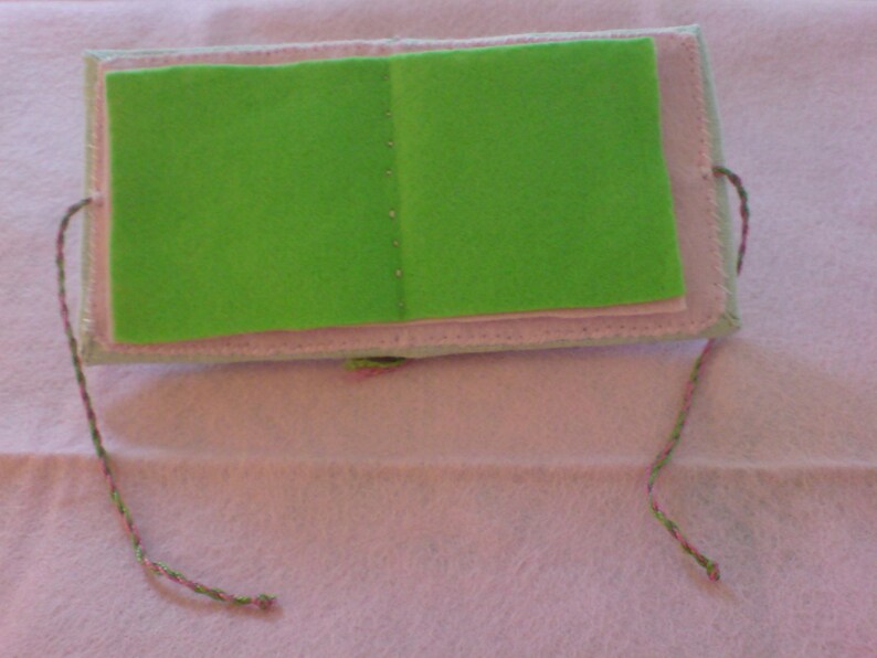 May include: A small green felt book with pink stitching and a pink and green string attached.