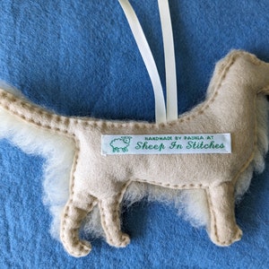 K&ouml;nnte beinhalten: Ein beigefarbener Filzhund-Anh&auml;nger mit einem wei&szlig;en, flauschigen Schwanz. Der Anh&auml;nger hat ein gr&uuml;nes Etikett mit der Aufschrift "Handmade by Paula at Sheep In Stitches".