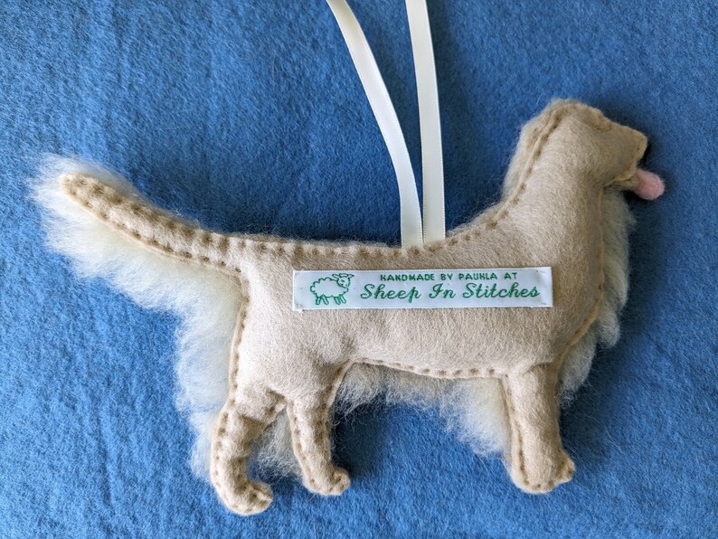 K&ouml;nnte beinhalten: Ein beigefarbener Filzhund-Anh&auml;nger mit einem wei&szlig;en, flauschigen Schwanz. Der Anh&auml;nger hat ein gr&uuml;nes Etikett mit der Aufschrift "Handmade by Paula at Sheep In Stitches".