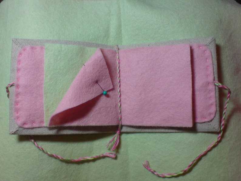 May include: A pink and green fabric book with a linen cover and a pin holding the pages together.
