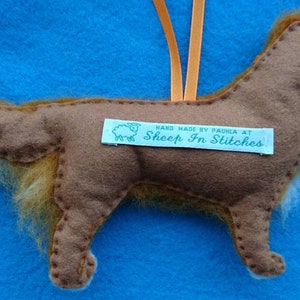 May include: A brown felt dog ornament with a fluffy tail. The ornament is hanging from a string and has a tag that reads "Hand Made by Paula at Sheep In Stitches".