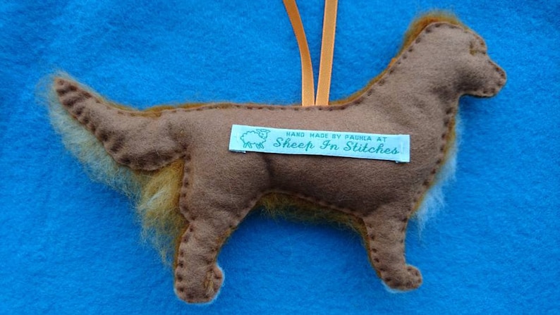 May include: A brown felt dog ornament with a fluffy tail. The ornament is hanging from a string and has a tag that reads "Hand Made by Paula at Sheep In Stitches".