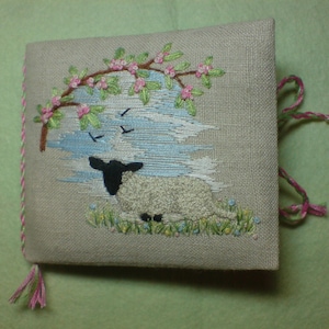 May include: A white lamb with black ears and hooves is embroidered on a beige linen fabric. The lamb is lying down in a field of green grass with pink flowers. A tree with pink flowers is embroidered in the upper left corner of the fabric.
