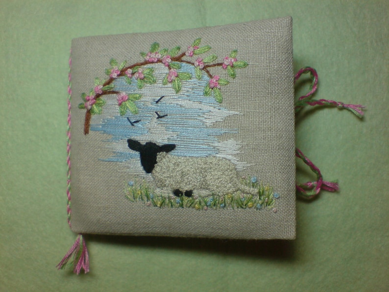 May include: A white lamb with black ears and hooves is embroidered on a beige linen fabric. The lamb is lying down in a field of green grass with pink flowers. A tree with pink flowers is embroidered in the upper left corner of the fabric.