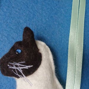 May include: A close-up of a handmade felt cat toy with a white body and brown face. The cat has a blue eye and white whiskers.