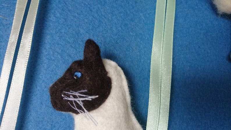 May include: A close-up of a handmade felt cat toy with a white body and brown face. The cat has a blue eye and white whiskers.