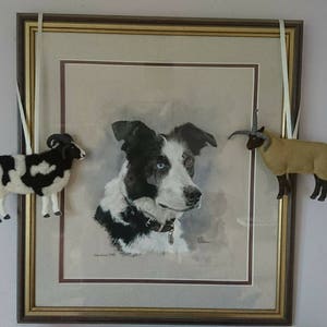 Puede incluir: Una pintura de acuarela enmarcada de un perro border collie blanco y negro. Dos peque&ntilde;os adornos de animales de fieltro, una oveja blanca y negra y una oveja marr&oacute;n, cuelgan del marco.
