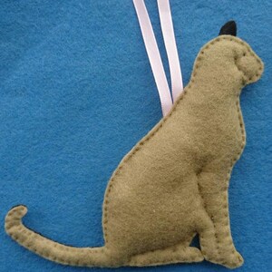 May include: A brown felt cat ornament with a pink ribbon hanging from the top. The cat is sitting with its tail curled around its body.