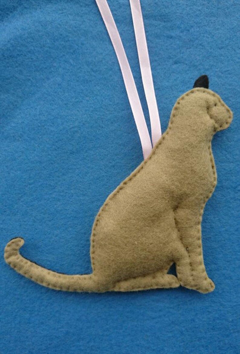 May include: A brown felt cat ornament with a pink ribbon hanging from the top. The cat is sitting with its tail curled around its body.
