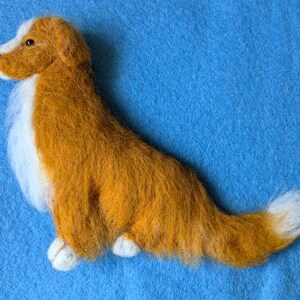 May include: A hand-felted dog sculpture, made of wool, in a reddish-brown color with white accents. The dog is lying on its side with its head turned to the left. The sculpture is on a blue background.
