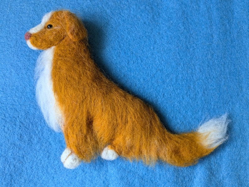 May include: A hand-felted dog sculpture, made of wool, in a reddish-brown color with white accents. The dog is lying on its side with its head turned to the left. The sculpture is on a blue background.