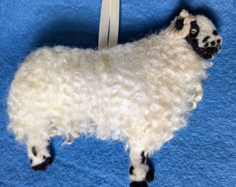 May include: A white sheep with black markings on its face and legs, made of wool felt. The sheep has curly white wool and a black face with a white patch on its forehead. The sheep is standing on a blue surface.