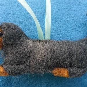 May include: A needle-felted ornament shaped like a dachshund dog with dark grey fur, tan accents on the face, paws, and tail, and a black nose. The dog has a light blue ribbon attached to the top for hanging, featuring a textured wool finish.