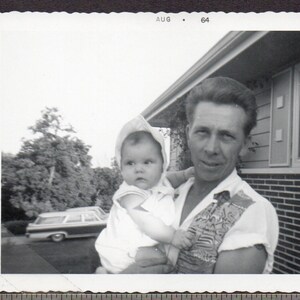 May include: A black and white photo of a man holding a baby in his arms. The man is wearing a patterned shirt and the baby is wearing a white dress. The photo was taken in August of 1964.