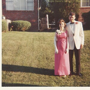 May include: A couple poses for a photo in front of a brick house. The woman is wearing a pink dress and white gloves. The man is wearing a white tuxedo jacket with a black bow tie. The photo is taken in a grassy area.