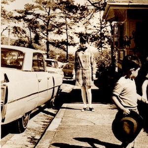 May include: A black and white vintage photograph of a woman standing in front of a white car. The woman is wearing a dress and a hat. There is a young boy standing in front of her, holding a hat. The photo was taken in front of a house.