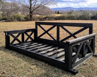 May include: A black wooden outdoor daybed with an X-shaped design on the back and sides. The daybed is made of dark-stained wood and sits on a grassy lawn. The frame has a slatted base.