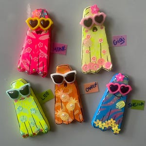 May include: A collection of five colorful ghost-shaped magnets, each with sunglasses and unique designs. The ghosts are in shades of pink, yellow, orange, and blue, decorated with floral and fruit patterns. Each magnet has a name tag.