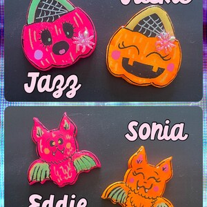 May include: Four Halloween-themed resin pins with cute faces. Two are orange pumpkin-shaped trick-or-treat bags with spiderwebs and glitter. Two are bats with pink and orange bodies and green wings.