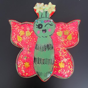 May include: A whimsical, handcrafted butterfly-shaped ornament. The butterfly features a green body with black stripes, red wings adorned with yellow glitter and floral accents. The butterfly has a smiling face and is topped with two yellow flowers. The background is black.