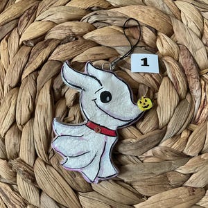 May include: A white Zero character ornament with a red collar and a small pumpkin on its nose. The ornament is on a woven background and has a tag with the number 1.