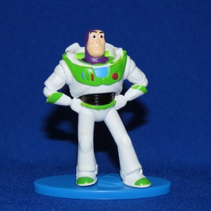 May include: A white and green plastic toy figure of Buzz Lightyear from the Toy Story franchise. The figure is standing with its arms crossed and has a purple helmet and a red button on its chest.