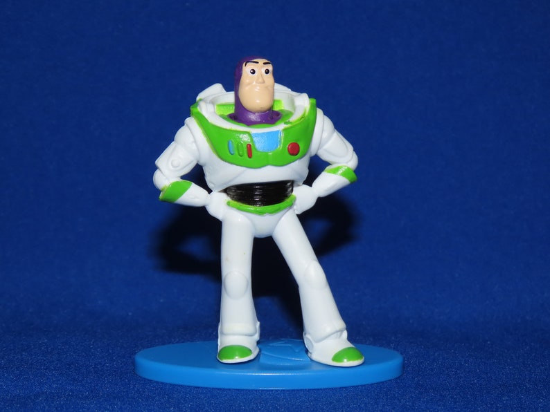 May include: A white and green plastic toy figure of Buzz Lightyear from the Toy Story franchise. The figure is standing with its arms crossed and has a purple helmet and a red button on its chest.