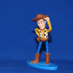 May include: A plastic toy figurine of Woody from the Toy Story franchise. Woody is wearing his signature brown cowboy hat, a yellow shirt with a red bandana, and blue jeans. He is holding his hat in his right hand and has a friendly expression on his face.