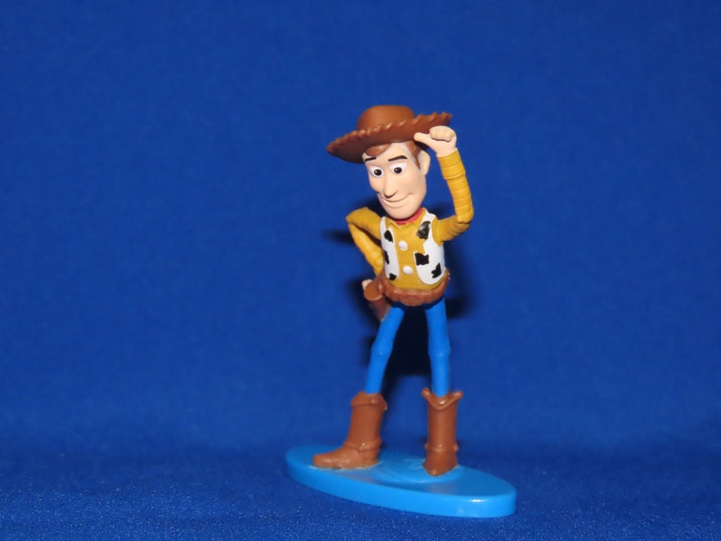 May include: A plastic toy figurine of Woody from the Toy Story franchise. Woody is wearing his signature brown cowboy hat, a yellow shirt with a red bandana, and blue jeans. He is holding his hat in his right hand and has a friendly expression on his face.