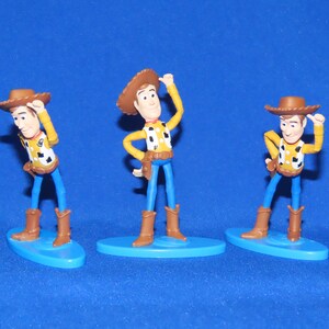 May include: Three plastic figurines of Woody from Toy Story. Each figurine is wearing a brown cowboy hat, a yellow shirt with a red bandana, and blue jeans. Each figurine is standing on a blue base.