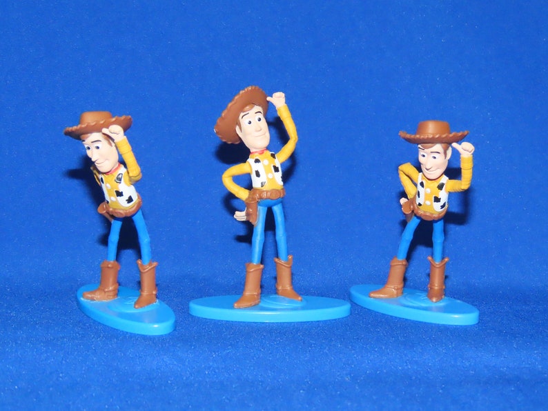 May include: Three plastic figurines of Woody from Toy Story. Each figurine is wearing a brown cowboy hat, a yellow shirt with a red bandana, and blue jeans. Each figurine is standing on a blue base.