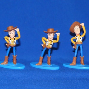 May include: Three plastic figurines of Woody from the Toy Story franchise. Each figurine is posed differently and stands on a blue base.
