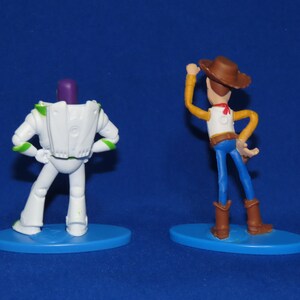 May include: Back view of two plastic action figures, Buzz Lightyear and Woody from the Toy Story franchise. Buzz Lightyear is white with green accents and a purple head. Woody is wearing a brown cowboy hat, a white shirt, a brown belt, blue jeans, and brown boots. Both figures are standing on blue bases.